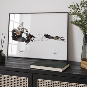 Op de afbeelding: Een zwart ingelijst afdruk met een aquarelkaart van Canada in zwart, goud en grijs. De kaart is verbonden door een lijn met twee harten. De tekst "George & Drew" staat onder de kaart met de datum "2 oktober 2015".