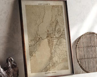 May include: A framed map print of Kelowna, Canada, in sepia tones. The map features detailed street and geographical outlines. The frame is a dark brown color. Coordinates are printed at the top. The text "KELOWNA, CANADA" is printed at the bottom.