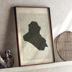 Może przedstawiać: Oprawiony wydruk przedstawiający mapę Iraku w ciemnej sylwetce, na tle tekstu w stylu vintage. Wydruk jest zamknięty w ciemnobrązowej drewnianej ramie, co dodaje klasycznego charakteru dziełu. Słowo "IRAQ" jest wydrukowane u góry.