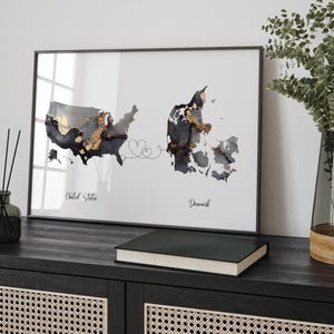 May include: A framed print of a map of the United States and Denmark connected by a line with hearts. The map is in black and white with gold accents. The text "United States" and "Denmark" is below the maps.