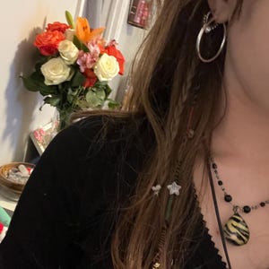 Może przedstawiać: Osoba z długimi brązowymi włosami w czarnej koszuli i naszyjnikiem z zawieszką w paski zebry. Osoba ma kolczyk w kształcie koła w lewym uchu. Na tle widać bukiet białych i pomarańczowych róż.
