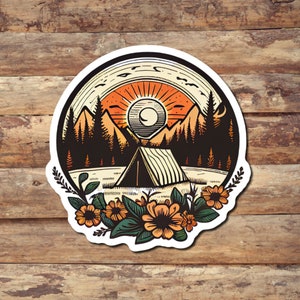 May include: A circular sticker featuring a camping scene. The design includes a tent, mountains, and a setting sun in shades of orange, brown, and green. Floral accents surround the bottom of the design, set against a white background.