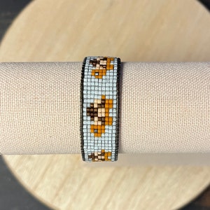 May include: A beaded bracelet with a repeating pattern of orange and brown floral designs on a light blue background. The bracelet is displayed on a beige cylindrical stand, showcasing the intricate beadwork and color palette.