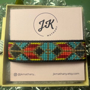 May include: A colourful beaded bracelet with a geometric pattern in shades of blue, red, yellow, and black. The bracelet is displayed in a box with the logo "JK Mathany" and the website address "jkmathany.etsy.com".