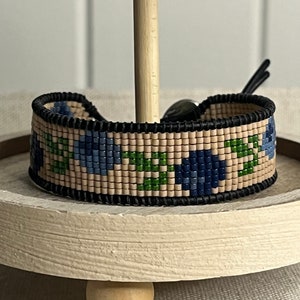 Könnte beinhalten: Ein Armband aus Perlen mit einem Blumenmuster in Blautönen, Grün und Beige. Das Armband ist aus kleinen, runden Perlen gefertigt und hat einen schwarzen Lederverschluss.