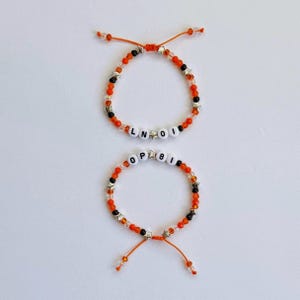 May include: Two adjustable bracelets made with orange, black, and clear beads, interspersed with small silver star-shaped charms. The top bracelet features white letter beads spelling LN OI, while the bottom bracelet features white letter beads spelling OP 8I.