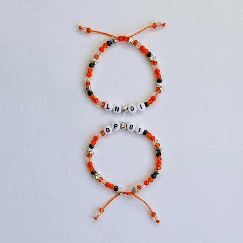 May include: Two adjustable bracelets made with orange, black, and clear beads, interspersed with small silver star-shaped charms. The top bracelet features white letter beads spelling LN OI, while the bottom bracelet features white letter beads spelling OP 8I.