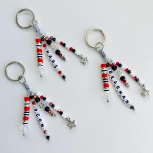 Op de afbeelding: Sleutelhangers met kralenaccenten in rood, wit, zwart en zilver. Elke sleutelhanger heeft een zilveren ring, een doorzichtige kristallen kraal en een sterbedel. De sleutelhangers hebben namen uitgeschreven in witte letterkralen.