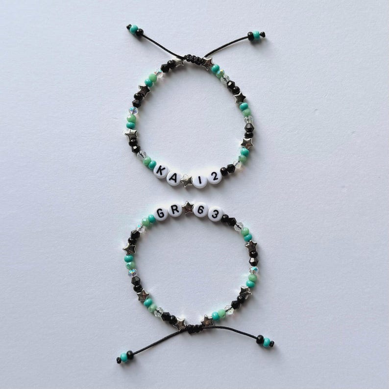 May include: Two adjustable bracelets featuring black, turquoise, and clear beads with silver-toned star accents. The top bracelet displays the text KA 12, while the bottom bracelet displays the text GR 63 on white letter beads.