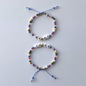 May include: Two adjustable bracelets featuring blue, red, white, and clear beads interspersed with small gold-coloured stars. One bracelet displays the letters MV and the numbers 03, while the other displays the letters IH and the numbers 06.
