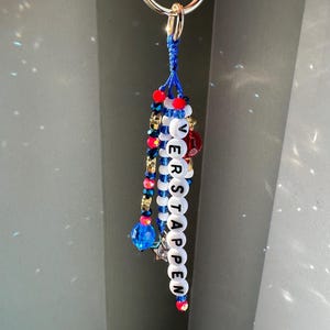 Pu&ograve; includere: Un portachiavi con un anello argentato e un design di perline blu, rosse e bianche. La parola "VERSTAPPEN" &egrave; scritta con perline di lettere bianche. Perline e ciondoli aggiuntivi in blu, rosso e oro completano il design.