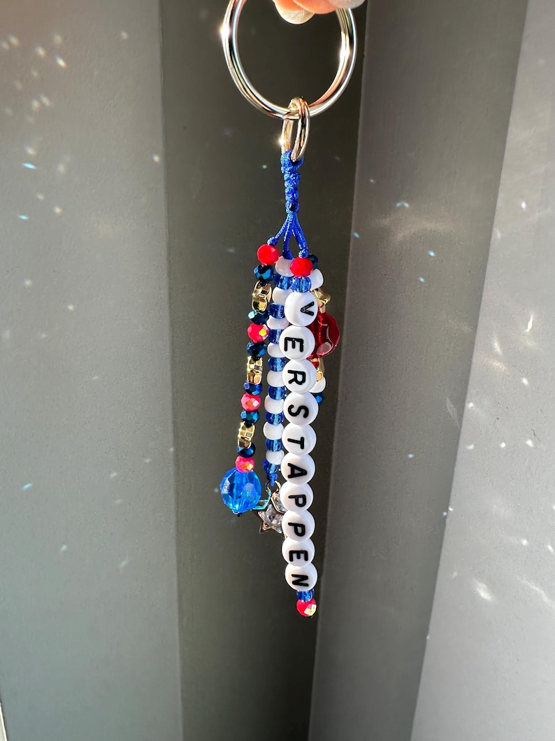 Pu&ograve; includere: Un portachiavi con un anello argentato e un design di perline blu, rosse e bianche. La parola "VERSTAPPEN" &egrave; scritta con perline di lettere bianche. Perline e ciondoli aggiuntivi in blu, rosso e oro completano il design.