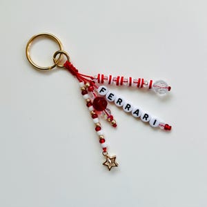 May include: A gold-colored keyring with a red and white beaded charm. The charm features the word "FERRARI" spelled out in white letter beads, along with a gold star charm and red and clear beads.