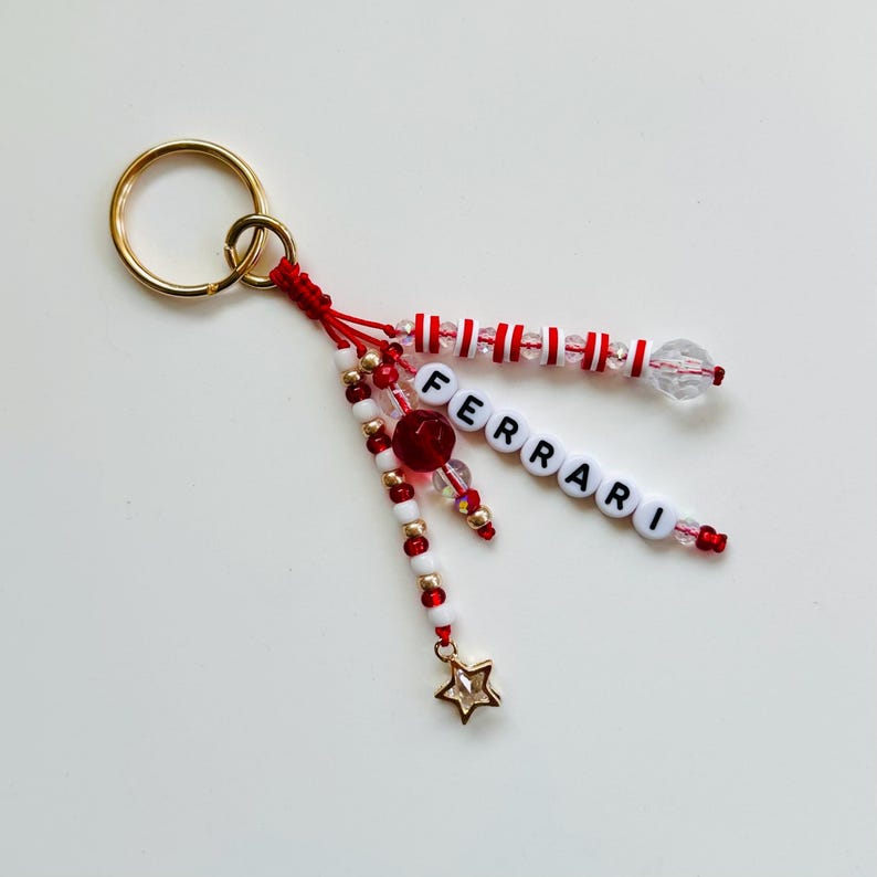 May include: A gold-colored keyring with a red and white beaded charm. The charm features the word "FERRARI" spelled out in white letter beads, along with a gold star charm and red and clear beads.