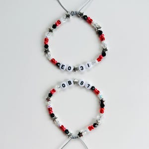 May include: Two adjustable beaded bracelets featuring red, black, and clear beads interspersed with silver star-shaped charms. One bracelet displays the text CEO 31, while the other displays the text OB 87, both using white letter and number beads.