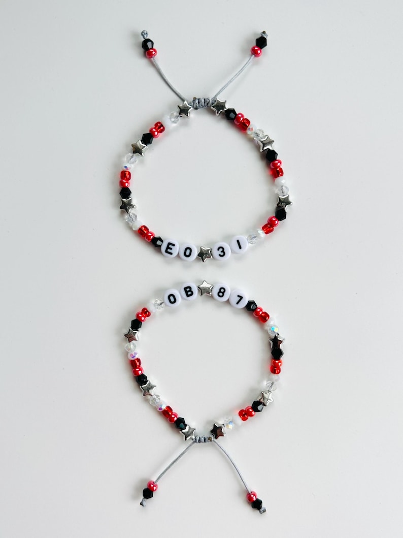 May include: Two adjustable beaded bracelets featuring red, black, and clear beads interspersed with silver star-shaped charms. One bracelet displays the text CEO 31, while the other displays the text OB 87, both using white letter and number beads.