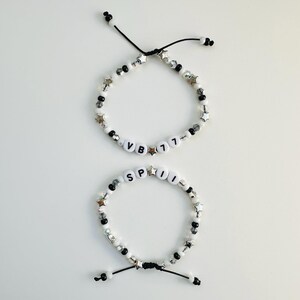 May include: Two adjustable bracelets featuring black and white beads, silver-toned star charms, and clear faceted beads. One bracelet displays the text VB 77, while the other displays the text SPII. Both bracelets are secured with black cord ties.