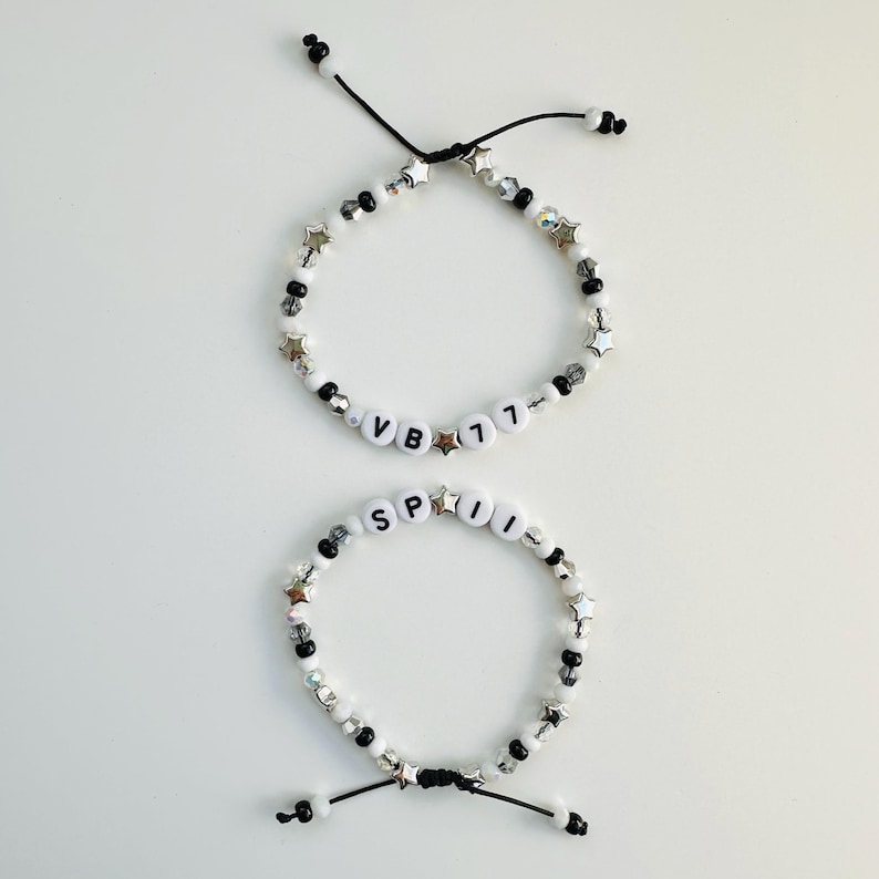 May include: Two adjustable bracelets featuring black and white beads, silver-toned star charms, and clear faceted beads. One bracelet displays the text VB 77, while the other displays the text SPII. Both bracelets are secured with black cord ties.