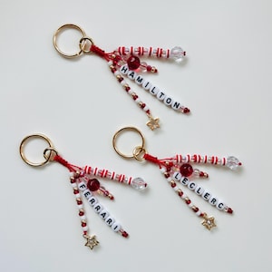 May include: Two keychains with gold rings, red string, and beaded accents. One keychain says "HAMILTON" and the other says "LECLERC". Both have gold star charms and red, white, and clear beads.