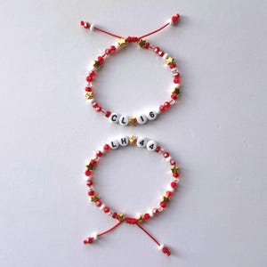 May include: Two adjustable bracelets made with red and clear beads, small gold star-shaped beads, and white letter beads. The top bracelet features the text CL 16, while the bottom bracelet displays the text LH 44. Both have red cord ends.