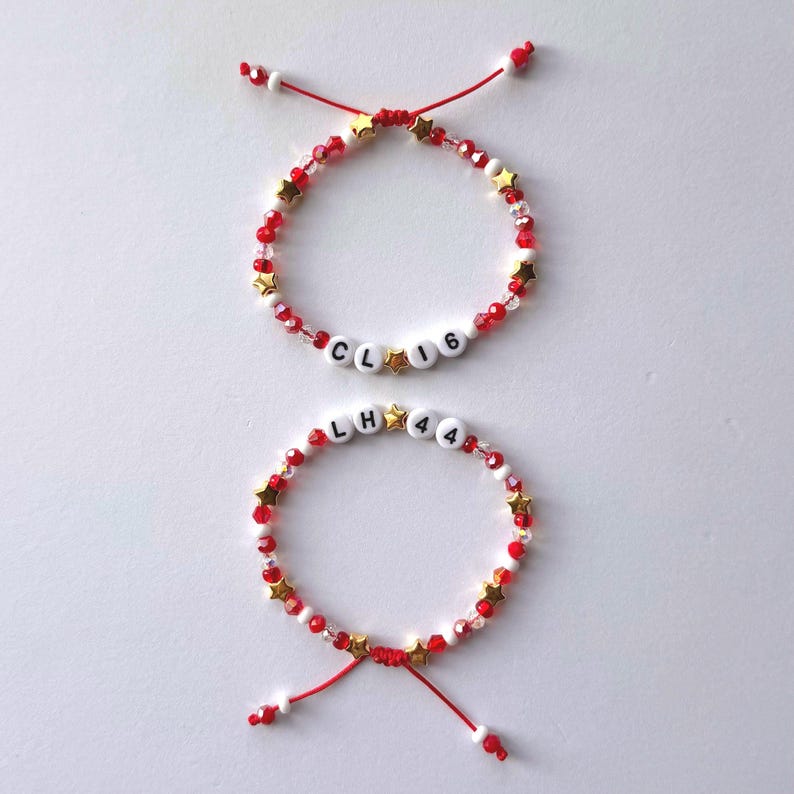 May include: Two adjustable bracelets made with red and clear beads, small gold star-shaped beads, and white letter beads. The top bracelet features the text CL 16, while the bottom bracelet displays the text LH 44. Both have red cord ends.