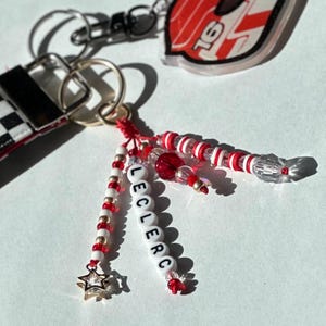 May include: A collection of keychains featuring a racing car design and beaded accents. One keychain spells out "LECLERC" with white letter beads. The keychains have red, white, and gold beads, and a star charm.