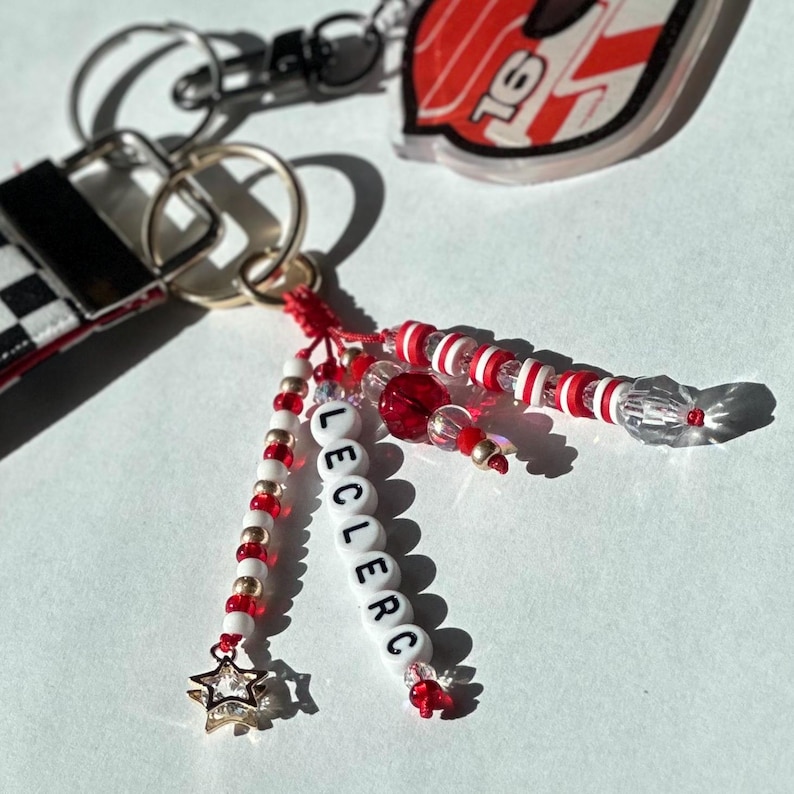 May include: A collection of keychains featuring a racing car design and beaded accents. One keychain spells out "LECLERC" with white letter beads. The keychains have red, white, and gold beads, and a star charm.