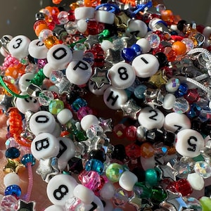 May include: A collection of colourful beaded bracelets featuring white round beads with black letters and numbers, interspersed with metallic silver star-shaped beads and various translucent, faceted, and solid-coloured plastic beads.