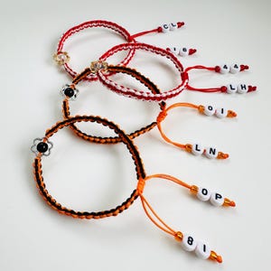 May include: Handmade friendship bracelets in red and orange, with white detailing. Each bracelet has a flower charm and personalised letter beads. The bracelets are displayed against a white background.