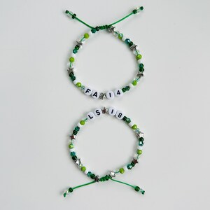 May include: Two adjustable bracelets featuring green and white beads, silver star-shaped charms, and white letter beads. The top bracelet displays the text FA 14, while the bottom bracelet displays the text LS 18. Both bracelets have green cord ends.