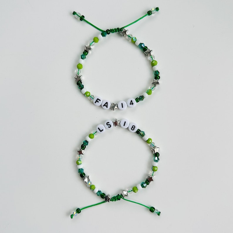 May include: Two adjustable bracelets featuring green and white beads, silver star-shaped charms, and white letter beads. The top bracelet displays the text FA 14, while the bottom bracelet displays the text LS 18. Both bracelets have green cord ends.