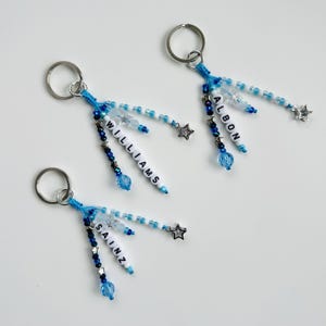 May include: Three keyrings with silver rings and blue beaded details. Each keyring features a cluster of beads in shades of blue and white, with a star charm and a name spelled out in letter beads. The names are "WILLIAMS", "ALBON", and "SAINZ".