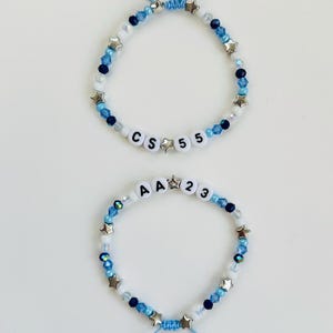 May include: Two adjustable bracelets featuring blue, white, and navy beads interspersed with silver-toned star charms. The top bracelet displays the letters CS and the number 55, while the bottom bracelet displays the letters AA and the number 23.