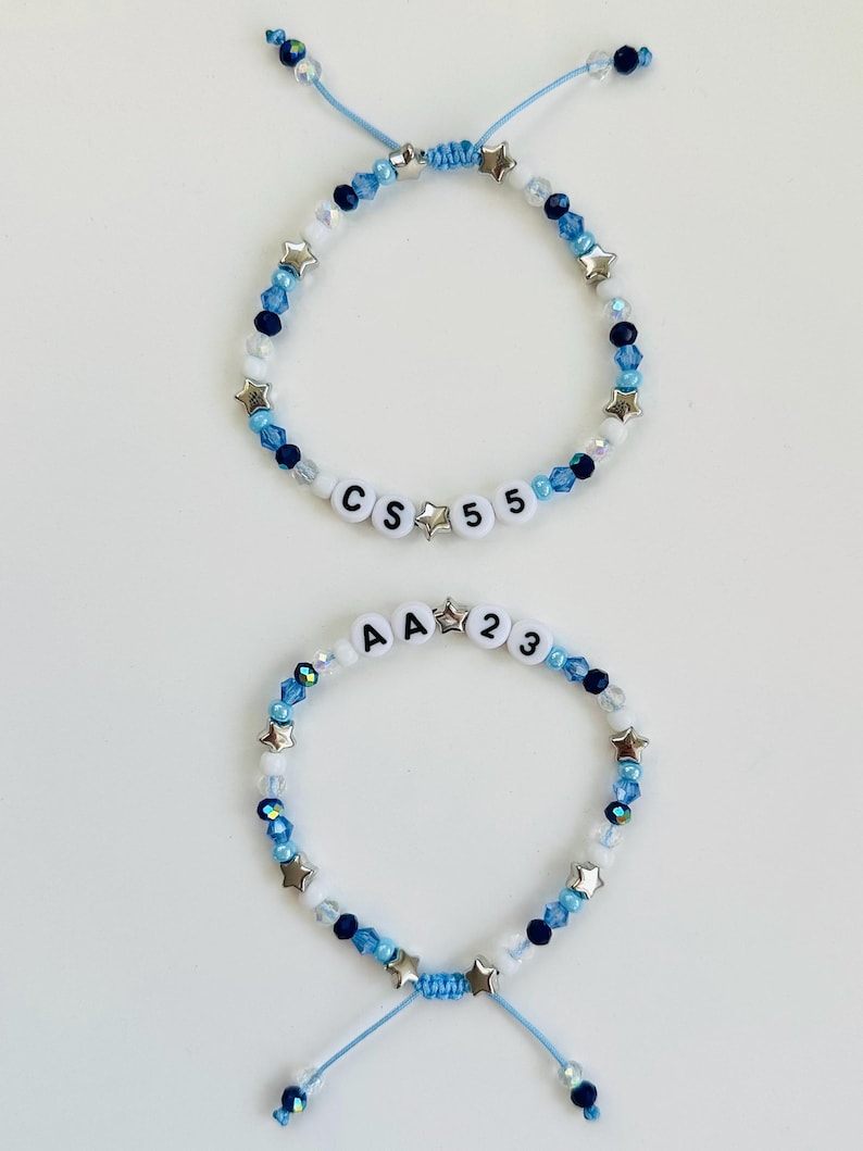 May include: Two adjustable bracelets featuring blue, white, and navy beads interspersed with silver-toned star charms. The top bracelet displays the letters CS and the number 55, while the bottom bracelet displays the letters AA and the number 23.