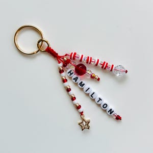 May include: A handmade keychain with gold-colored ring and red cord. The keychain features white beads, red beads, and gold beads. The word "HAMILTON" is spelled out with white letter beads. A gold star charm hangs from the bottom.
