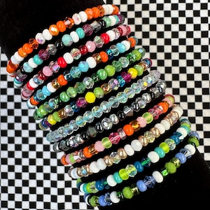 May include: A collection of eleven elastic bracelets featuring a variety of colourful beads. The beads include opaque white, translucent faceted crystals, and solid-coloured spheres in shades of orange, blue, green, pink, yellow, and metallic tones.