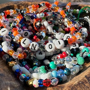 May include: A collection of colourful beaded bracelets featuring white alphabet and number beads, faceted translucent crystals, and metallic star-shaped spacers. The bracelets are strung on elastic cord and display various letters and numbers.