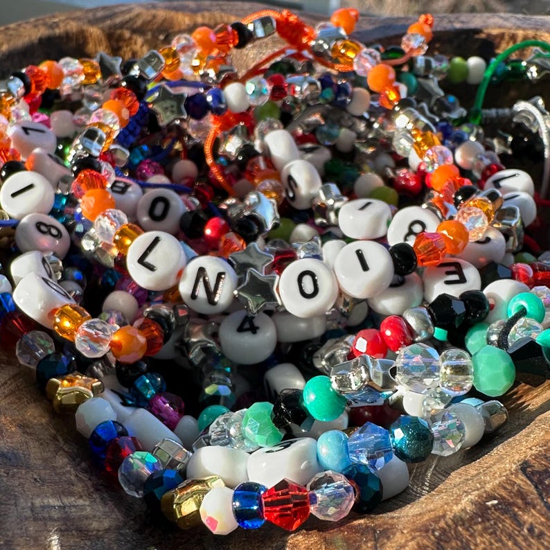 May include: A collection of colourful beaded bracelets featuring white alphabet and number beads, faceted translucent crystals, and metallic star-shaped spacers. The bracelets are strung on elastic cord and display various letters and numbers.