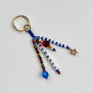 Pu&ograve; includere: Un portachiavi fatto a mano con un anello dorato e dettagli di perline blu, rosse e bianche. Il portachiavi presenta la parola "REDBULL" scritta con perline bianche, un ciondolo a forma di stella dorata e una perlina a forma di rosa rossa.