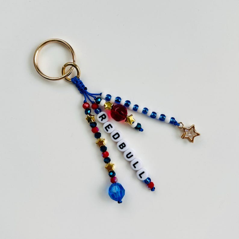 Pu&ograve; includere: Un portachiavi fatto a mano con un anello dorato e dettagli di perline blu, rosse e bianche. Il portachiavi presenta la parola "REDBULL" scritta con perline bianche, un ciondolo a forma di stella dorata e una perlina a forma di rosa rossa.