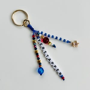 Pu&ograve; includere: Un portachiavi con un anello in metallo dorato collegato a tre fili di perline. I fili includono perline blu, rosse e bianche, ciondoli a forma di stella dorata e perline con lettere bianche che compongono il nome VERSTAPPEN in verticale.
