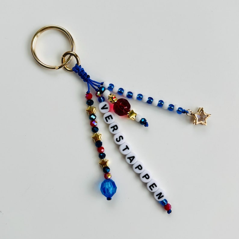 Pu&ograve; includere: Un portachiavi con un anello in metallo dorato collegato a tre fili di perline. I fili includono perline blu, rosse e bianche, ciondoli a forma di stella dorata e perline con lettere bianche che compongono il nome VERSTAPPEN in verticale.