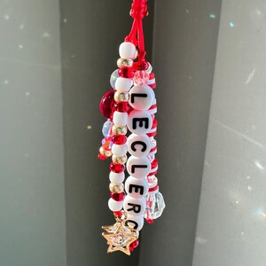 May include: A handmade keychain with red, white, and gold beads spelling "LECLERC". The keychain features a gold star charm and a red cord loop. Additional clear and red beads are interspersed throughout the design.