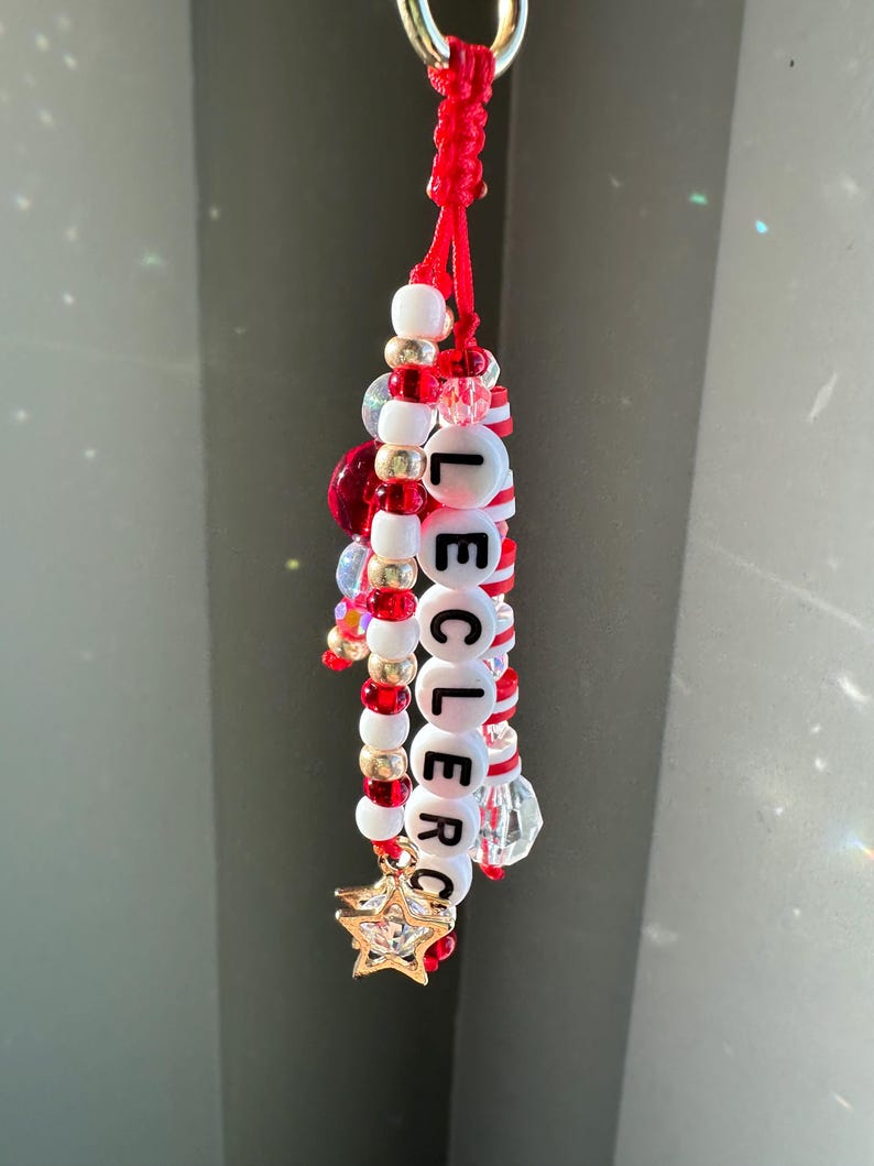 May include: A handmade keychain with red, white, and gold beads spelling "LECLERC". The keychain features a gold star charm and a red cord loop. Additional clear and red beads are interspersed throughout the design.