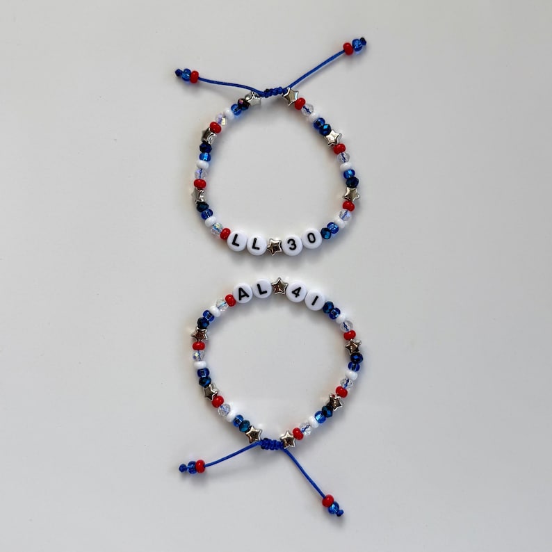 May include: Two adjustable bracelets featuring red, white, and blue beads, silver star-shaped spacers, and white letter beads. One bracelet displays the text LL 30, while the other displays the text AL 41, both strung on blue cord.