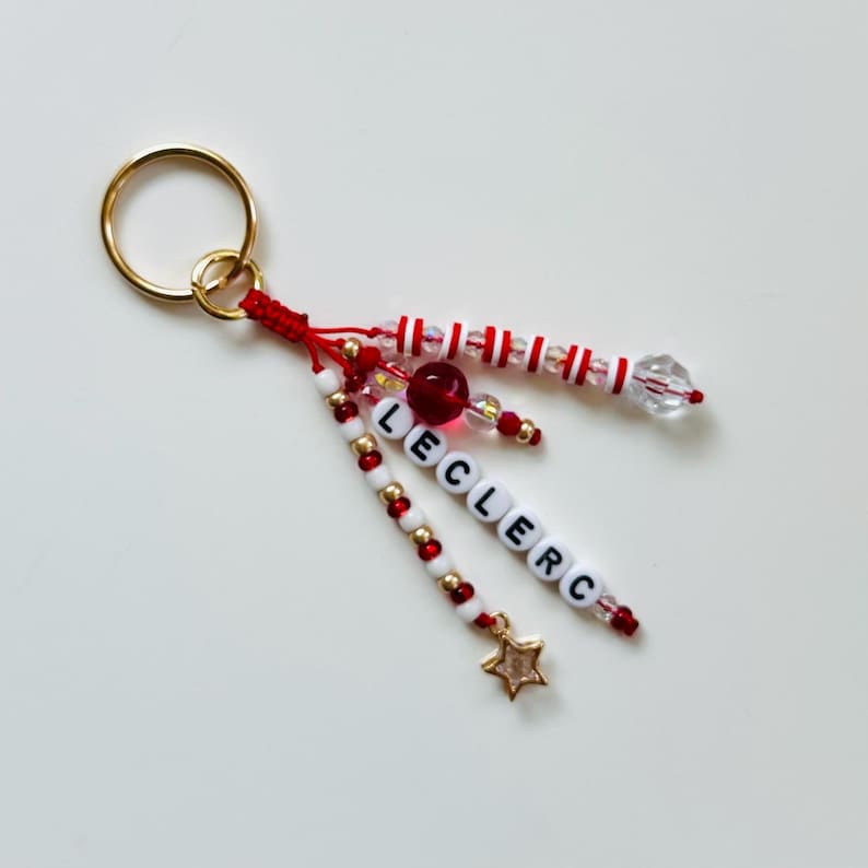 May include: A handmade keychain with a gold ring and red string. The keychain features white and red beads, gold beads, and the name "LECLERC" spelled out in white letter beads. A gold star charm hangs from the bottom.