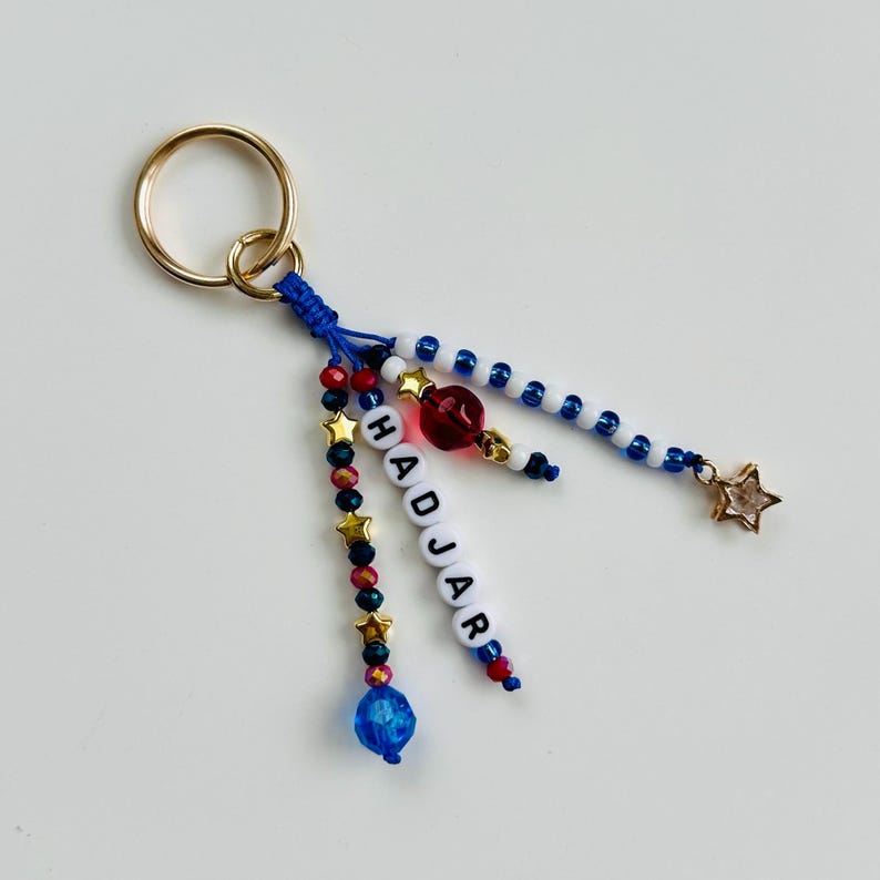 Pu&ograve; includere: Un portachiavi con un anello in metallo dorato attaccato a cordini blu. I fili sono decorati con perline rosse, blu e bianche, ciondoli a forma di stella dorata e perline con lettere bianche che compongono HADJAR, con un ciondolo a stella trasparente.