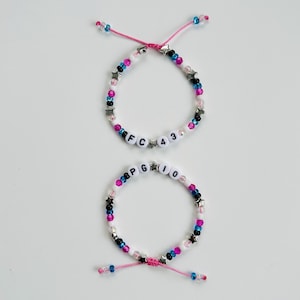 May include: Two adjustable beaded bracelets featuring pink, blue, black, and white beads with silver star accents. The top bracelet displays the text FC 43, while the bottom bracelet displays the text PG 10, both using white letter beads.