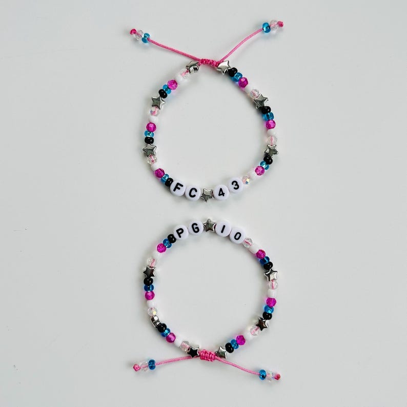 May include: Two adjustable beaded bracelets featuring pink, blue, black, and white beads with silver star accents. The top bracelet displays the text FC 43, while the bottom bracelet displays the text PG 10, both using white letter beads.