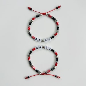 May include: Two adjustable bracelets featuring red and black beads, silver star-shaped spacers, and white letter beads. The top bracelet displays the text GB 05, while the bottom bracelet displays the text NH 27, both strung on red cord.